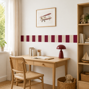 Burgundy and almond striped fabric wallpaper border above a wooden desk with a burgundy lamp and framed airplane print.